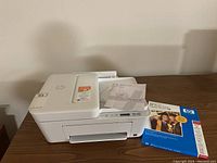 White HP DeskJet 4100e printer on a wooden surface with manuals atop and a package of photo paper beside it.
