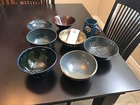 Top-down view of all seven bowls arranged in a group with one tall mug, showing various glaze colors and patterns.
