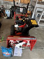 Front view showing red auger housing, white metal auger blades, manual crank and included operator manual and fuel jug on floor