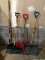 Photo of three snow shovels standing upright against a concrete block wall: one with a large black blade and wooden handle; one with red blade and red D-grip handle; one with grey blade and blue D-grip handle