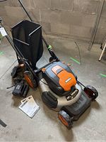 Front angle view of mower with bag, batteries, charger, and manual on concrete floor