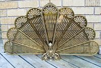 Full front view of brass fireplace screen showing peacock fan design and central gargoyle figure.