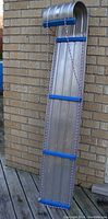 Full view of the aluminum toboggan standing upright against a brick wall showing length, cross braces, and rope handles.