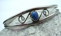 Close-up oblique view of the silver-tone adjustable cuff bracelet showing oval blue stone centerpiece with serrated bezel and swirl metalwork details on either side, set on a gray stone background.