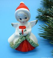 Front view of the tiny vintage Christmas angel figurine showing the angel's face, red Santa hat, white robe, and holding a gift with holly detail.