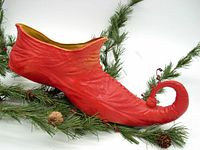 Side view of a red vintage rubber elf shoe with curly toe and pom-pom, decorated with artificial greenery around it.