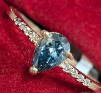 Front view of ring showing pear-shaped blue diamond and pave-set shoulder diamonds on red velvet