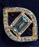 Top-down view of ring showing emerald-cut blue center stone and surrounding round diamonds on dark background