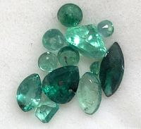 Close-up of multiple green emerald gemstones on a white surface showing varying shapes and inclusions.