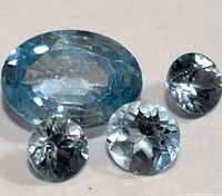 Four blue zircon gemstones including one large oval cut and three smaller round cuts shown on a pale background.