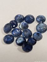 Close-up photo of thirteen genuine round cut sapphire gemstones with visible facets and deep blue color on a white background.