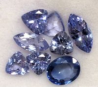 Close-up photo of eight faceted tanzanite gemstones showing various cuts and deep blue to violet colors on a light background.