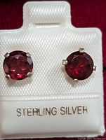 Close-up front view of the stud earrings showing round garnet stones in sterling silver prong settings on a white holder marked 'STERLING SILVER'.