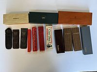 Photo showing 15 pen cases and pouches arranged on white surface, some leather, some wood or cardboard boxes, with visible brand names like Parker, Sheaffer, Easywriter.