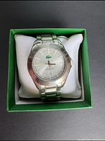 Front view of Lacoste men's watch showing silver dial, Lacoste logo, and date window in green watch box.