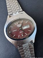 Close-up of watch face showing Sicura brand, automatic movement, day-date function, burgundy striped dial.