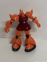 Front view of the articulated Zaku II action figure showing the full body with orange-red armor and black details on knees and feet.