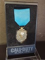 Front view of the silver-toned Call of Duty: Black Ops collectible medal with blue ribbon inside a black display case with foam insert.