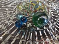 Vintage glass marbles assortment with various colors and patterns in a cut glass dish, showing mostly yellow, green, and blue marbles with swirls and bubbles inside.