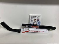Three items on white background: NHL Slapshot Wii game case standing, two hockey stick controller attachments placed horizontally. Black controller with NHL Slapshot branding, white controller with Team Canada branding.