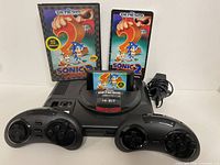 Front view of Sega Genesis Flashback console with Sonic 2 cartridge inserted, two wireless controllers, power adapter, original game case and manual behind console.