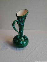 Front angled view of the vintage green ceramic vase on a white surface showing the elongated neck, flared top, and curved handle with light blue polka dot glaze accents.