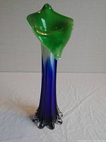 Front angled view of tall glass vase with green flared top and blue to black gradient stem ending in clear petal-like base on white surface.