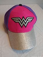 Front view of a vibrant pink baseball cap with metallic silver Wonder Woman logo and silver glitter brim on a white surface.