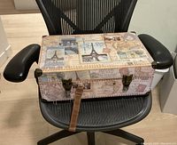 Decorative travel case with travel-themed stamped design placed on a chair, showing broken handle and latches.