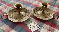 Two brass candle holders on a checkered cloth, showing the floral engraved design and handles.