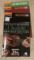 Front covers of four ukulele instruction books: Hal Leonard Method Books 1 and 2, Easy Songs for Ukulele, and Absolute Beginners Omnibus Edition by Steven Sproat.