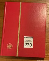 Photo of the red hardcover stamp album closed, showing its cover with a gold stripe and circular emblem.