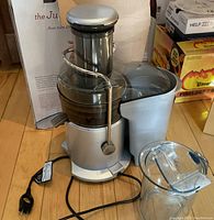 Front view of Breville Juice Fountain Plus juicer with attached clear juice pitcher, power cord visible, placed on wood floor near original box.