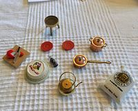 All miniature dollhouse kitchen items including cookware, tableware, small food, and kitchen accessories displayed on white textured cloth. Items include copper pots, teapot, red plates, cake, pepper shaker, flour sack, cutting board with cheese and knife, and a small grill.