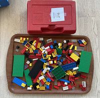 Red LEGO case placed on floor beside several assorted LEGO pieces of various colors and sizes on a wooden tray.