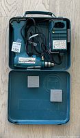 Open hard plastic carrying case containing Makita cordless drill, charger with attached cord