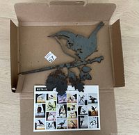 Metal bird wall art sculpture lying in original box, showing the bird perched on a branch with leaves and the included instruction sheet with multiple bird images.