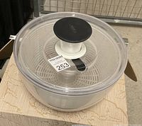 Clear plastic OXO vegetable/salad spinner showing top black spinning knob on transparent lid with OXO logo and attached inner basket.