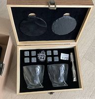 Photo showing open wooden gift box with 2 crystal whiskey glasses, 8 whiskey stones, pair of metal tongs, and 2 round coasters inside black velvet lining.