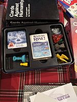 Photo showing Cards Against Humanity game pieces including cards, blue and yellow pawns, and colored dice in a plastic organizer.