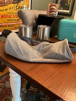 View of striped cloth wine carrier holding two stainless steel insulated wine cups with clear lids placed on a wooden table with background furniture.