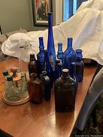 Photo showing 18 assorted colored glass bottles in blue, brown and clear, arranged on a table with a white covered background.