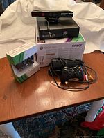 Xbox 360 console with Kinect sensor on top, boxed Kinect packaging, and one wireless controller outside the box along with AV composite cable displayed on wooden table.
