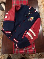 Front view of a kids varsity style jacket with navy wool body, red leather sleeves with white stripes, and a colorful patch on the chest.
