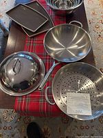 Overview of stainless steel wok with handle, strainer insert, lid, some ceramic trays, and nested mixing bowls on a table with a red plaid runner.