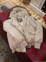 Photo showing an off white Roots down filled bomber jacket laid out on a table, highlighting the fur-lined hood, patch on sleeve, front pockets, and ribbed cuffs and hem.