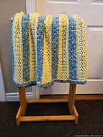 Crochet blanket draped over wooden rack, showing texture and colors of yellow, blue, and cream stripes.