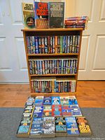 Photo showing wooden bookshelf with 4 shelves of softcover Clive Cussler novels plus 3 hardcover books on top and a display of 23 softcovers laid neatly in rows on floor.