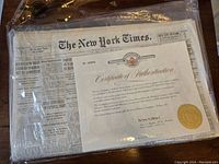 Front view showing folded The New York Times newspaper inside clear protective sleeve, certificate visible on top.
