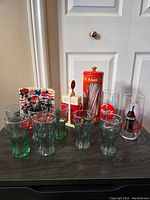 Overview of the Coca-Cola glassware and kitchen collectibles arranged on a dark surface, showing glasses, napkin holder, and various containers.